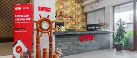 Een hotel receptie met een houten klok, een rode OYO app promotie stand en een modern receptiebalie met een logo.