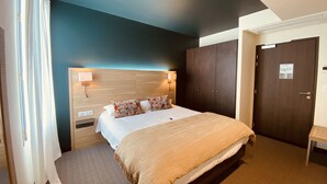Habitación de hotel con cama, cabecera de madera, mesita de noche, lámpara de pared, armario y puerta con un cartel.