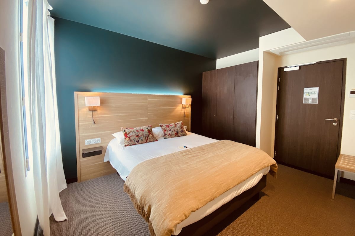 A hotel room with a bed, a wooden headboard, a nightstand, a wall-mounted lamp, a wardrobe, and a door with a sign.