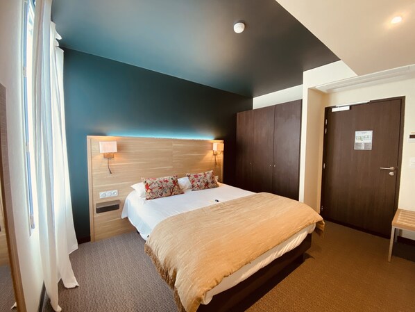 A hotel room with a bed, a wooden headboard, a nightstand, a wall-mounted lamp, a wardrobe, and a door with a sign. - Le Relais des Gourmands (Gramat)