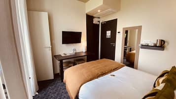 Habitación de hotel con cama, escritorio, televisor, espejo y estante con tetera.