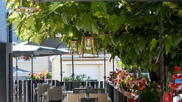 Un área de comedor al aire libre con mesas y sillas, cubierta por un toldo y rodeada de follaje y flores.