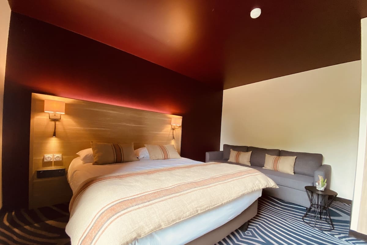 A modern hotel room with a large bed, a sofa, a small table, and a wall-mounted lamp.