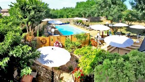 Zona de piscina con sillones de descanso y sombrillas, rodeada de exuberante vegetación y un muro de piedra.