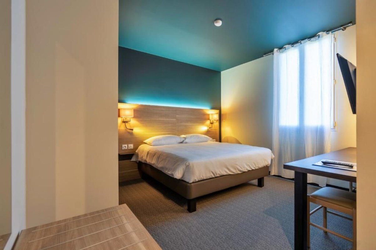 Una habitación de hotel con cama, lámparas de noche, escritorio y ventana con cortinas.