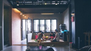 A group of people sitting on a bench in a modern interior space with a brick wall, large windows, and a ceiling fan.