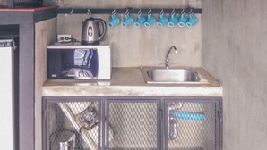 A compact kitchen with a microwave, kettle, and sink. There are potted plants on a shelf and a hanging rack with cups.