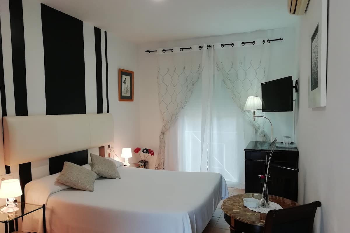 A hotel room with a bed, bedside lamp, a small table with a lamp, a chair, a desk with a lamp, a TV, and a window with curtains.