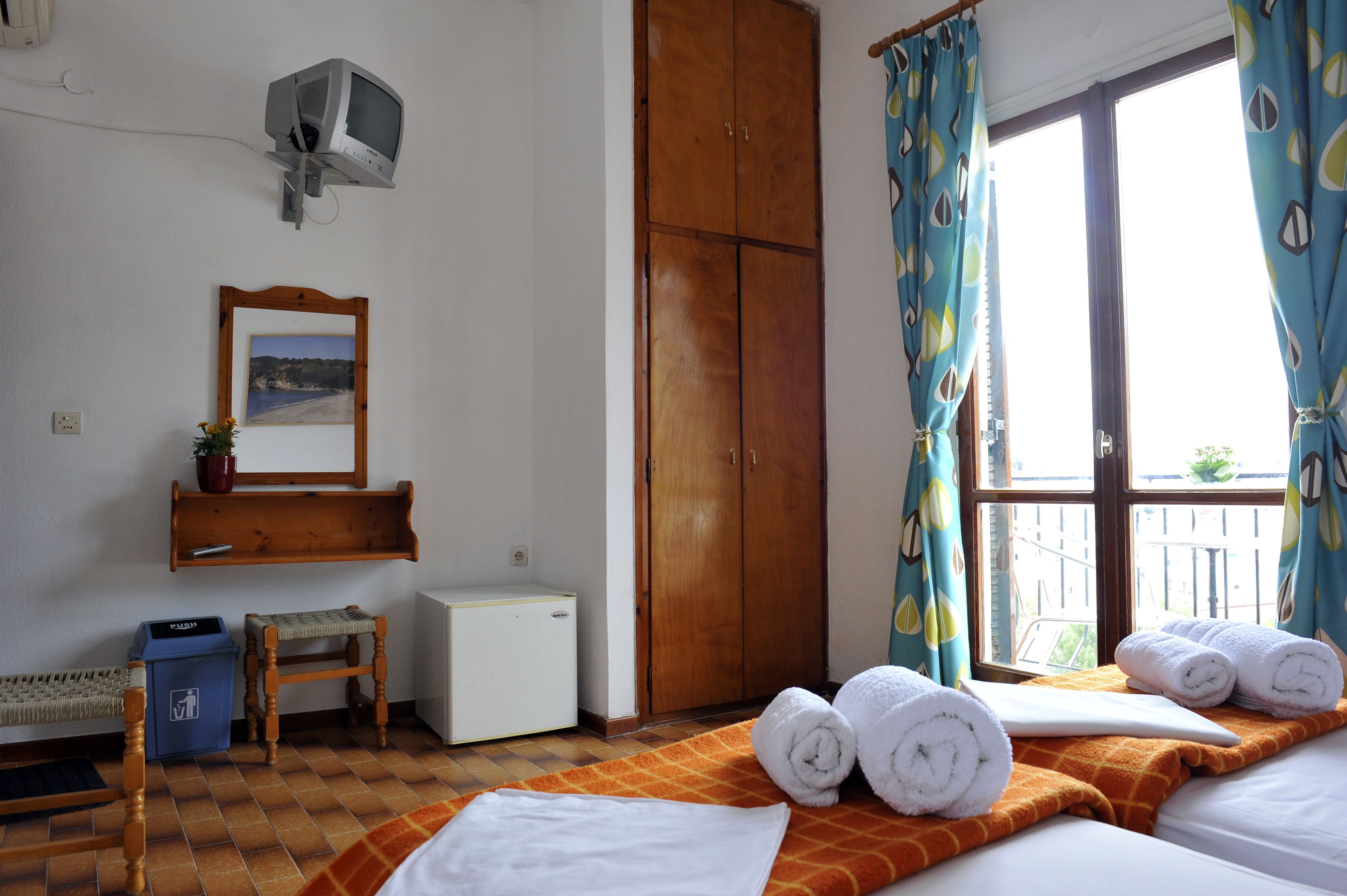 Quarto de hotel com uma cama, uma mesinha, uma cadeira, um guarda-roupa, um espelho e uma janela com cortinas.