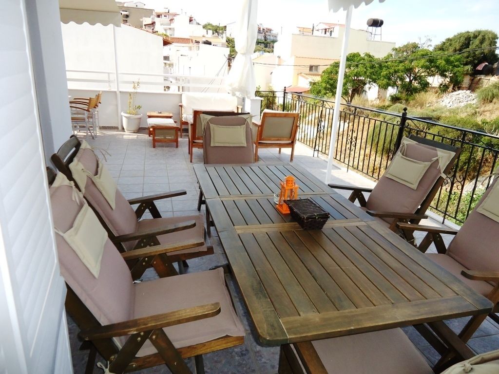 Une terrasse avec des tables et des chaises en bois, une petite valise orange et une vue sur les bâtiments et la végétation environnants.