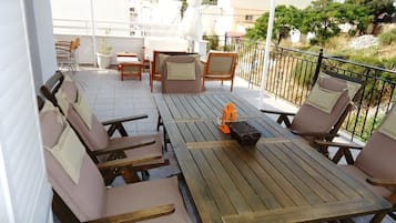Um terraço com mesas e cadeiras de madeira, uma mala laranja pequena e vista dos edifícios e vegetação ao redor.