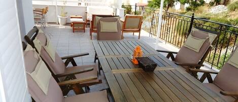 Um terraço com mesas e cadeiras de madeira, uma mala laranja pequena e vista dos edifícios e vegetação ao redor.
