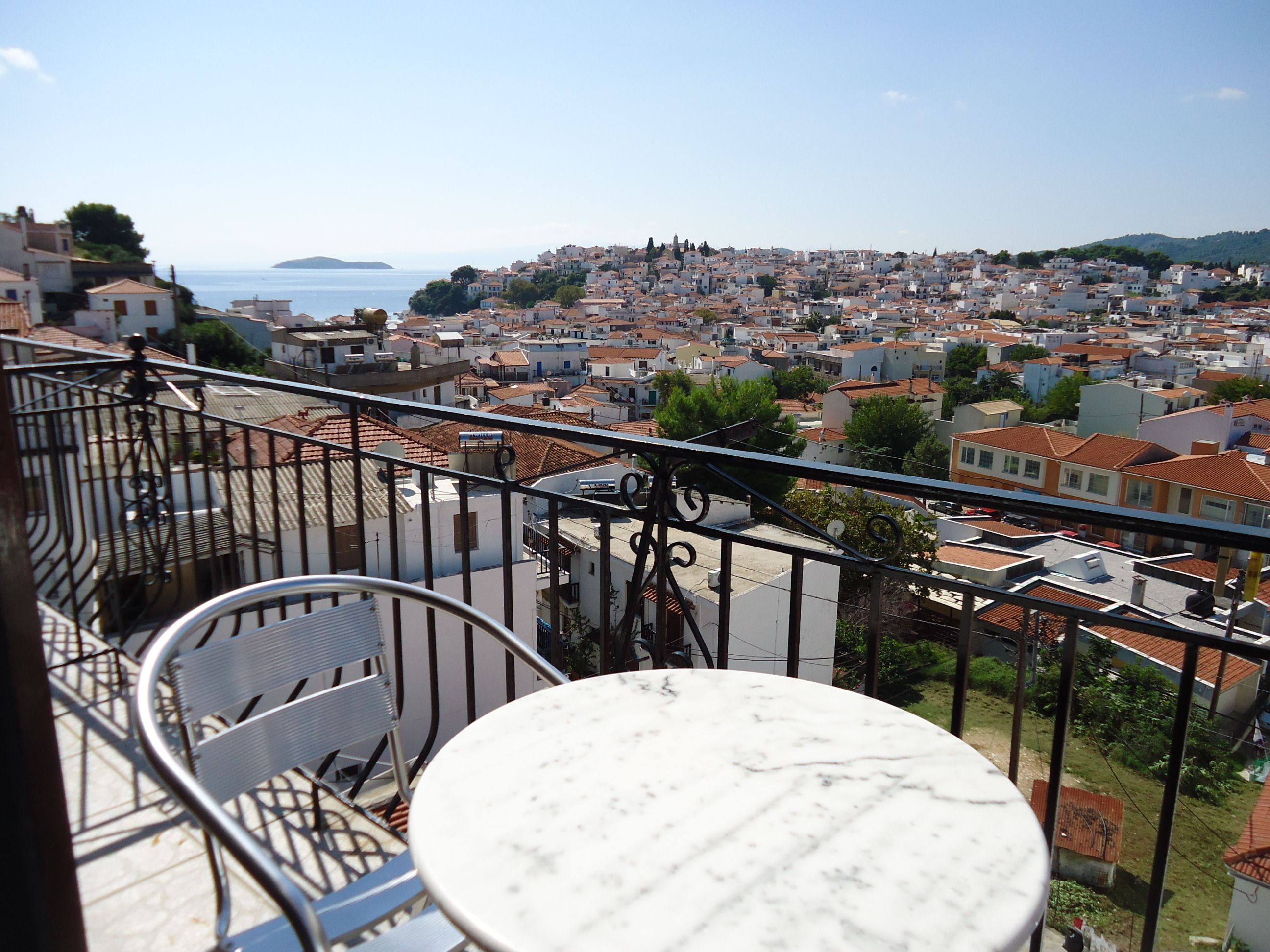 Un balcón con una mesa redonda y sillas metálicas, con vista a un pueblo costero de edificios con tejados de terracota y al mar.