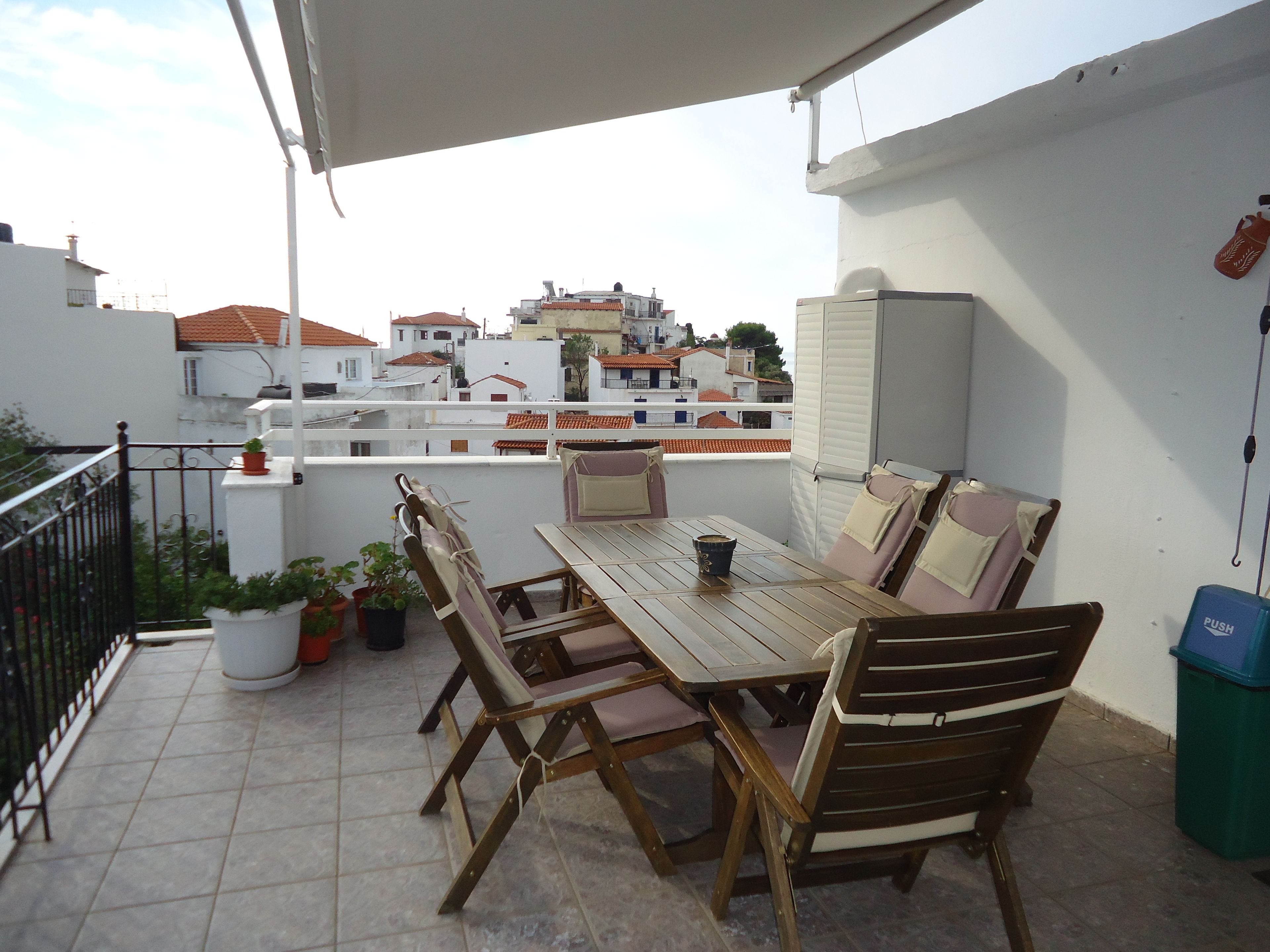 A terrace with a wooden table and chairs, potted plants, and a view of buildings in the background.
