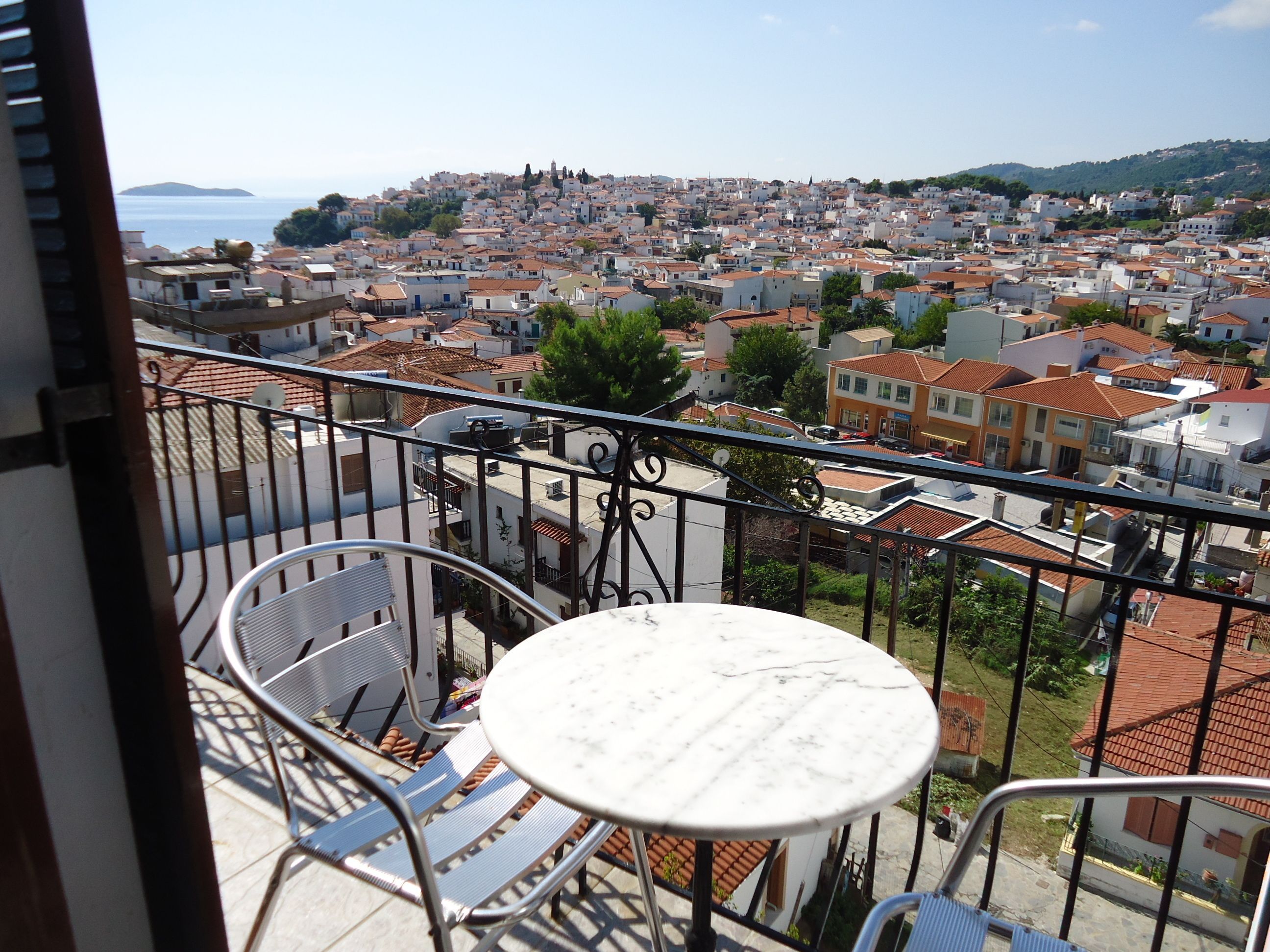 A balcony with a round table and two chairs, overlooking a densely packed town with red-roofed buildings and a distant view of the sea.