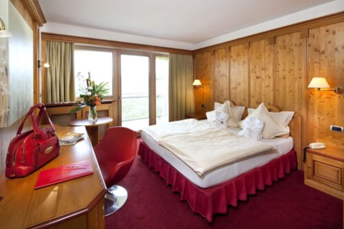 Habitación de hotel con pared de paneles de madera, una cama grande con ropa de cama blanca, una silla roja, un escritorio y un bolso rojo encima, y dos lámparas de noche.