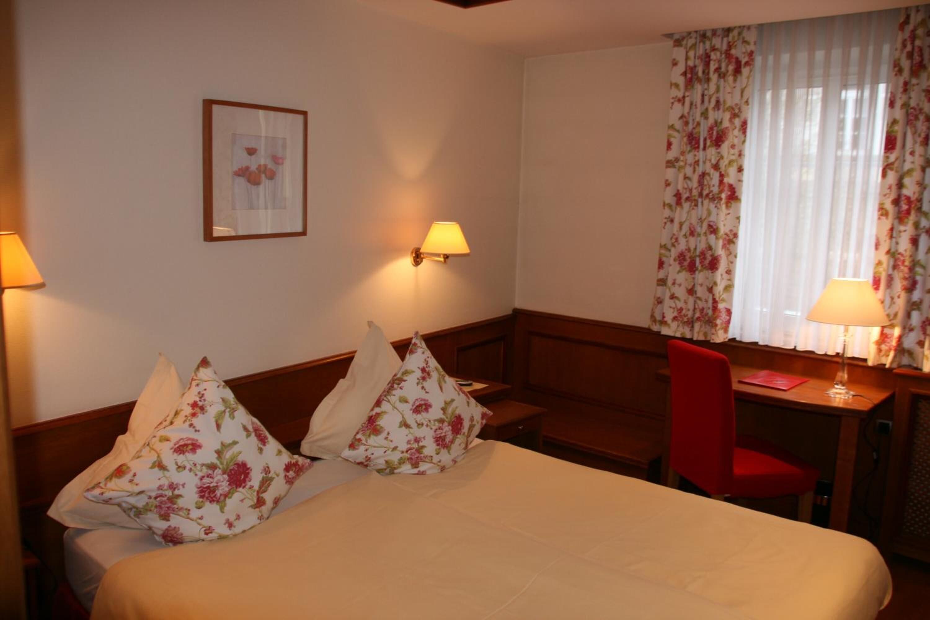 A hotel room with a bed, two pillows, a desk, a chair, a lamp, and a window with curtains.