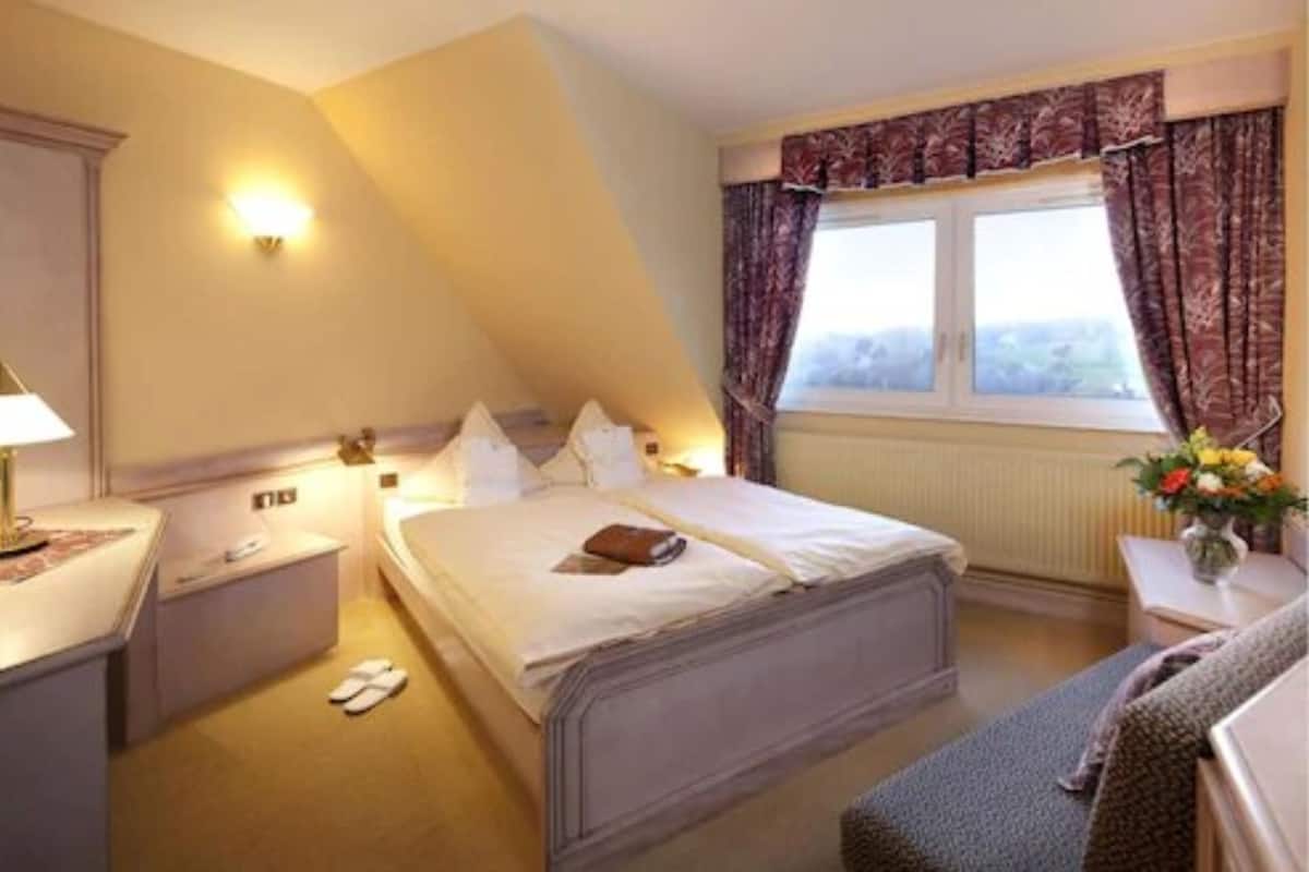 Habitación de hotel con una cama individual, mesitas de noche, una lámpara, un ventanal con cortinas y vistas al exterior.
