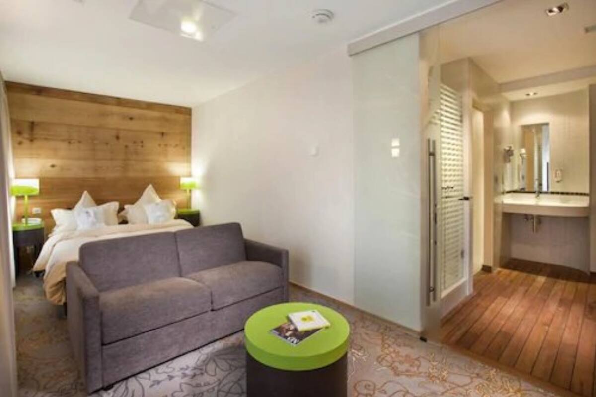 Habitación de hotel con cama, sofá gris, una mesita pequeña verde y baño con suelo de madera y lavamanos.
