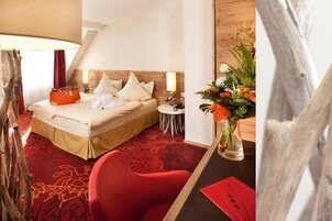 Una habitación de hotel con una cama, una silla roja, una mesita con un florero y una lámpara.