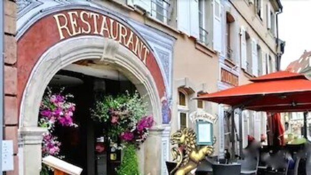 A restaurant with an ornate entrance, a statue of a mythical creature, and outdoor seating with a red awning. - SPA Hôtel Au Lion D'Or (La Petite-Pierre)
