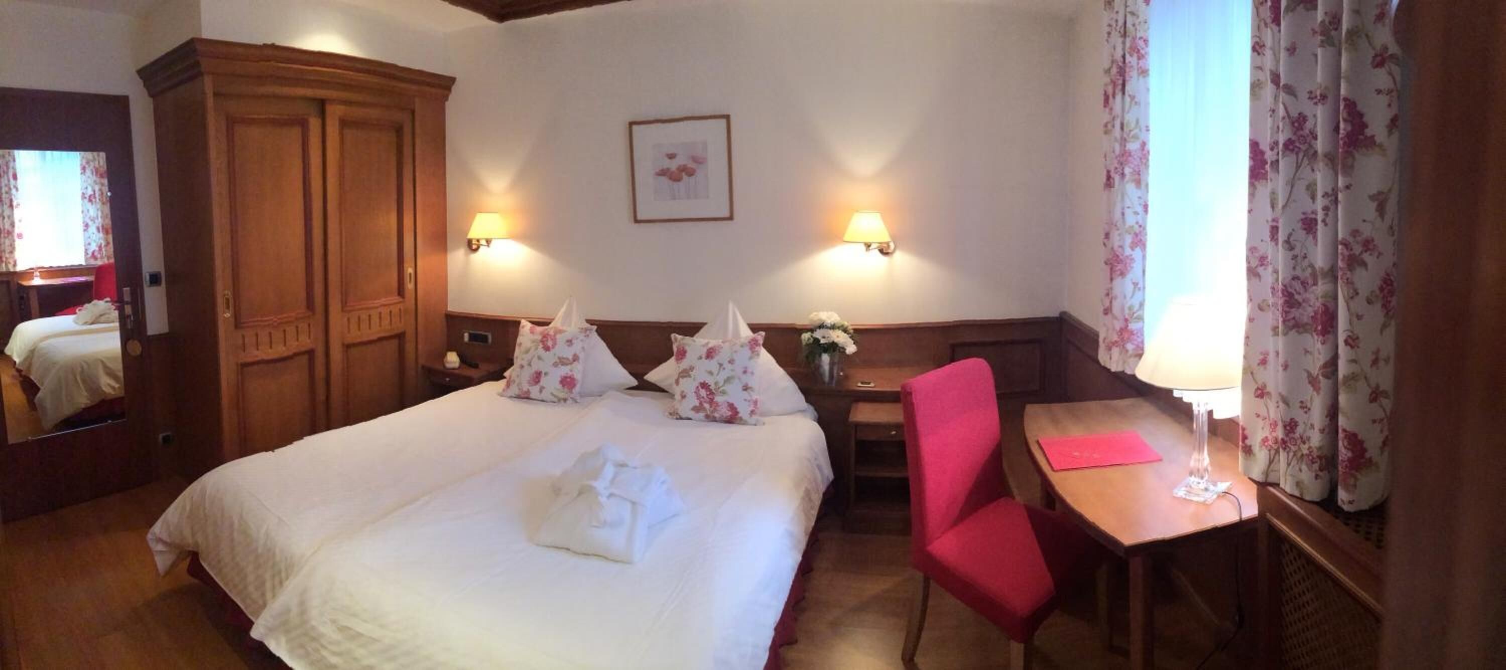 A hotel room with a large bed, two bedside lamps, a desk with a chair, and a wooden wardrobe.