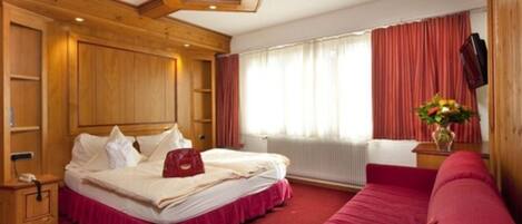 Ein Hotelzimmer mit einem hölzernen Kopfteil, einem Bett mit rotem Überwurf, einem roten Sofa, einem Nachttisch aus Holz und einem Fenster mit Vorhängen.