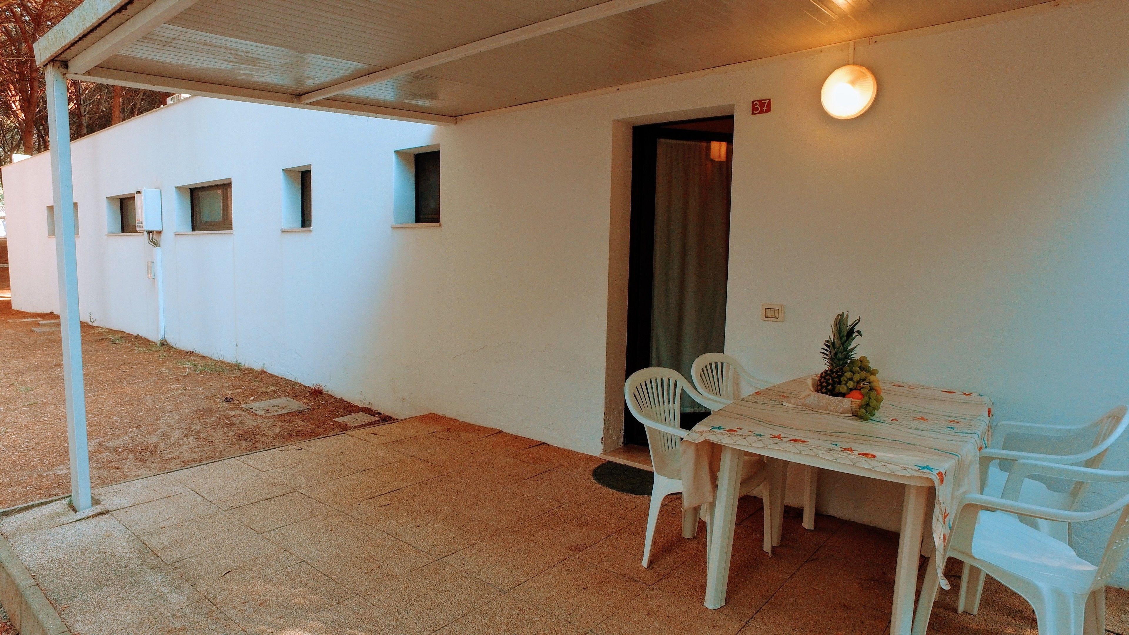 A covered outdoor dining area with a table and chairs, a small garden, and a building with numbered windows.