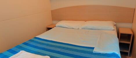 Una cama individual con colcha azul y blanca a rayas, dos almohadas blancas y cabecera de madera. Hay dos mesitas de noche de madera, una a cada lado de la cama.