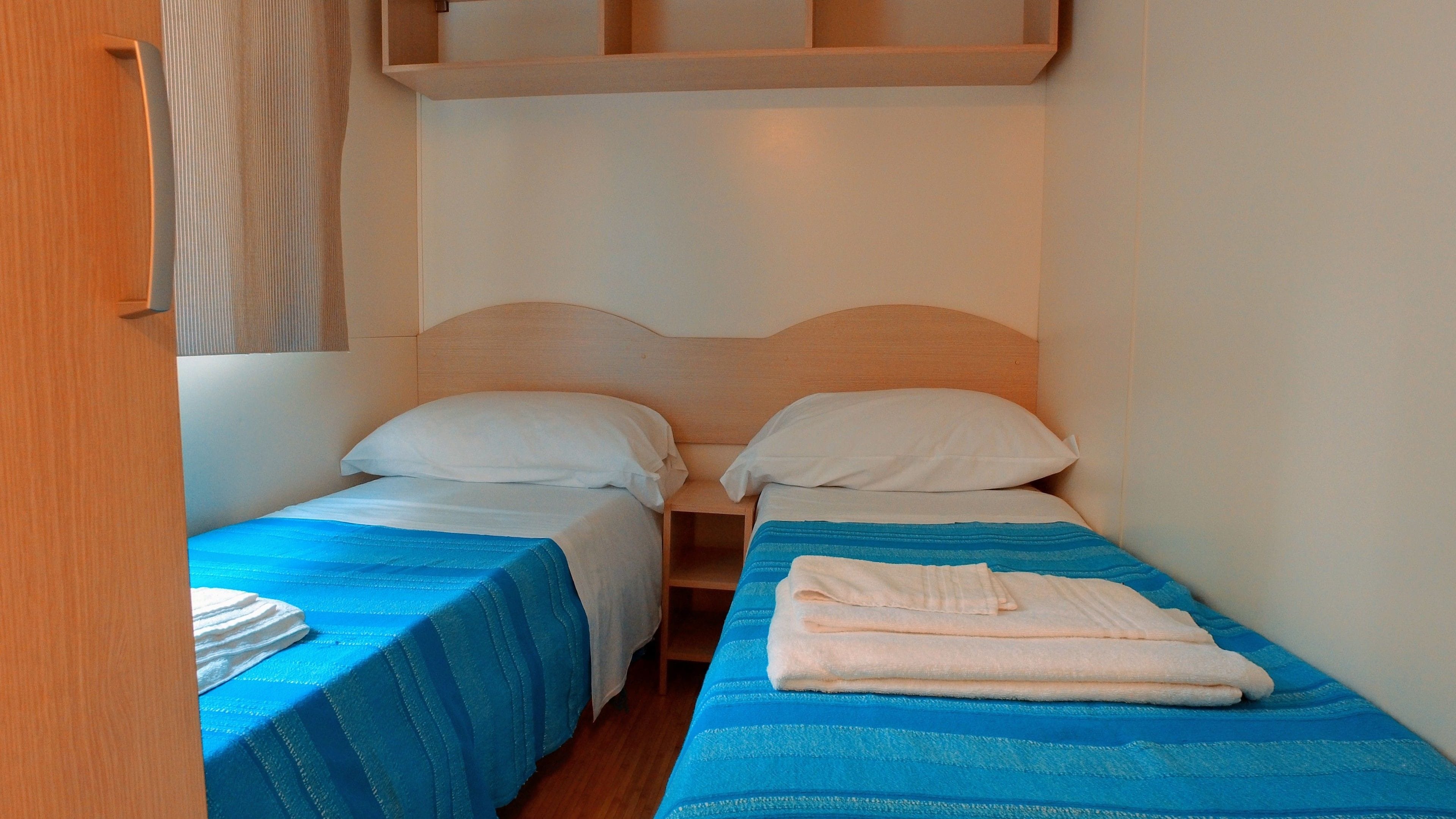 A small, compact room with two single beds, each with a blue bedspread and white pillows. There is a wooden headboard and a small shelf above the beds.