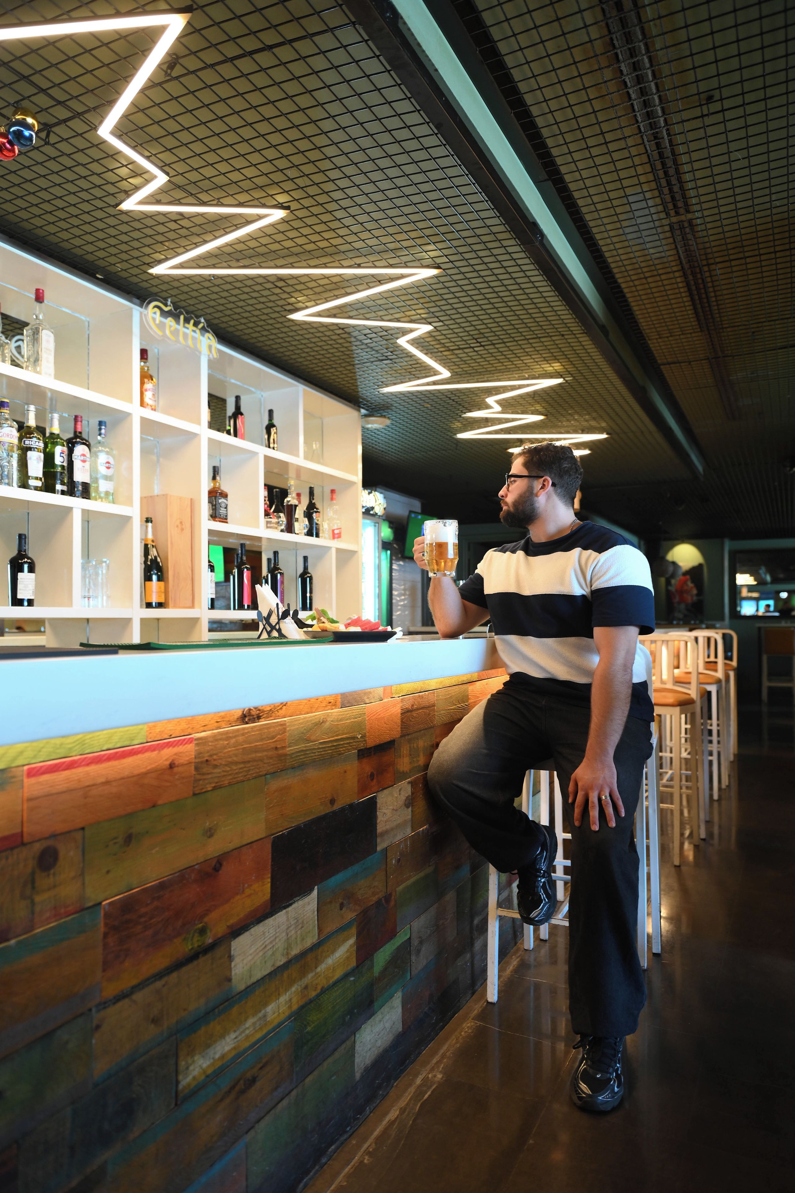 Una persona sentada en una barra, con una variedad de botellas y vasos en estantes al fondo.
