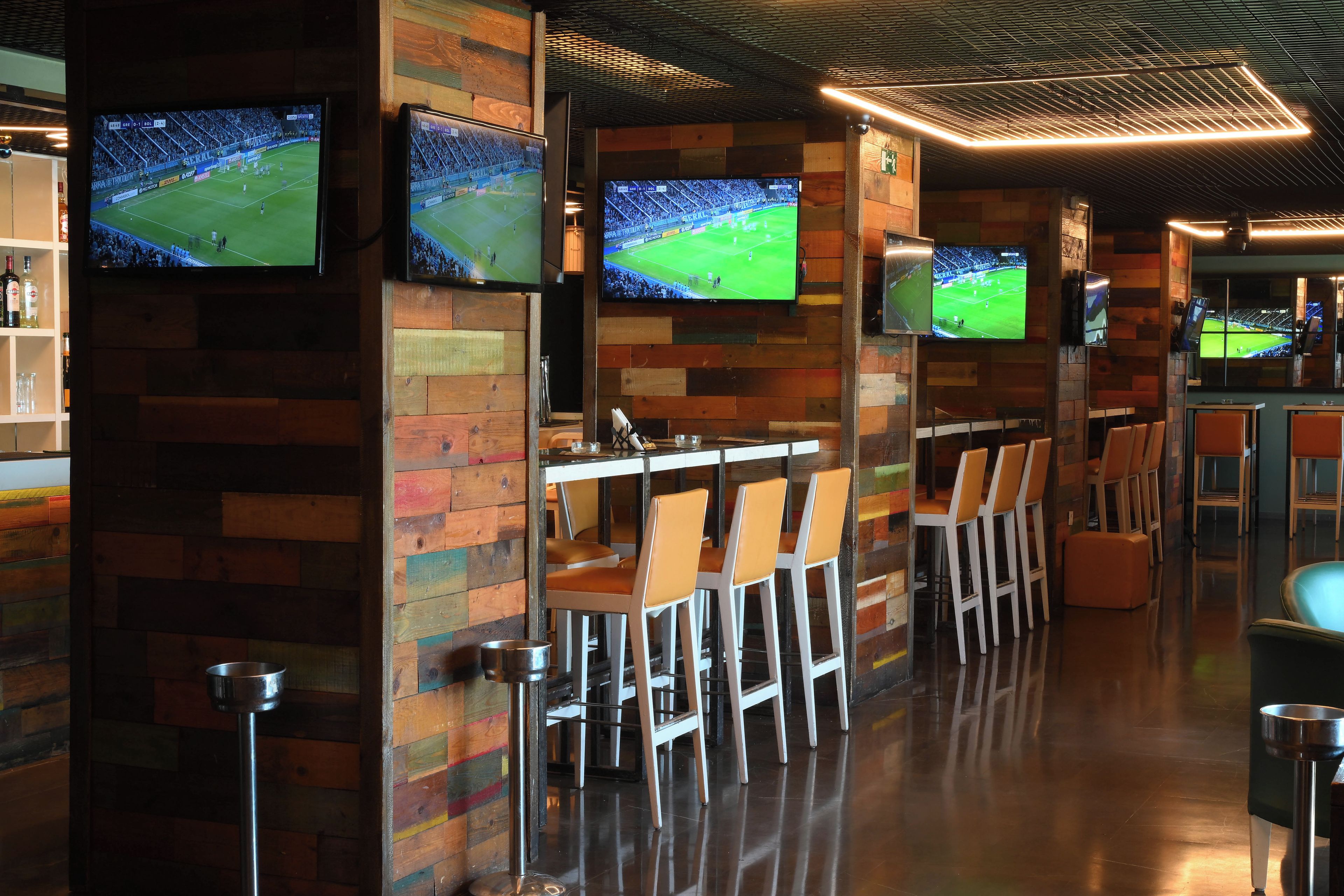 Eine Bar mit mehreren Bildschirmen, die Sportübertragungen zeigen, Holzwänden und Barhocker.