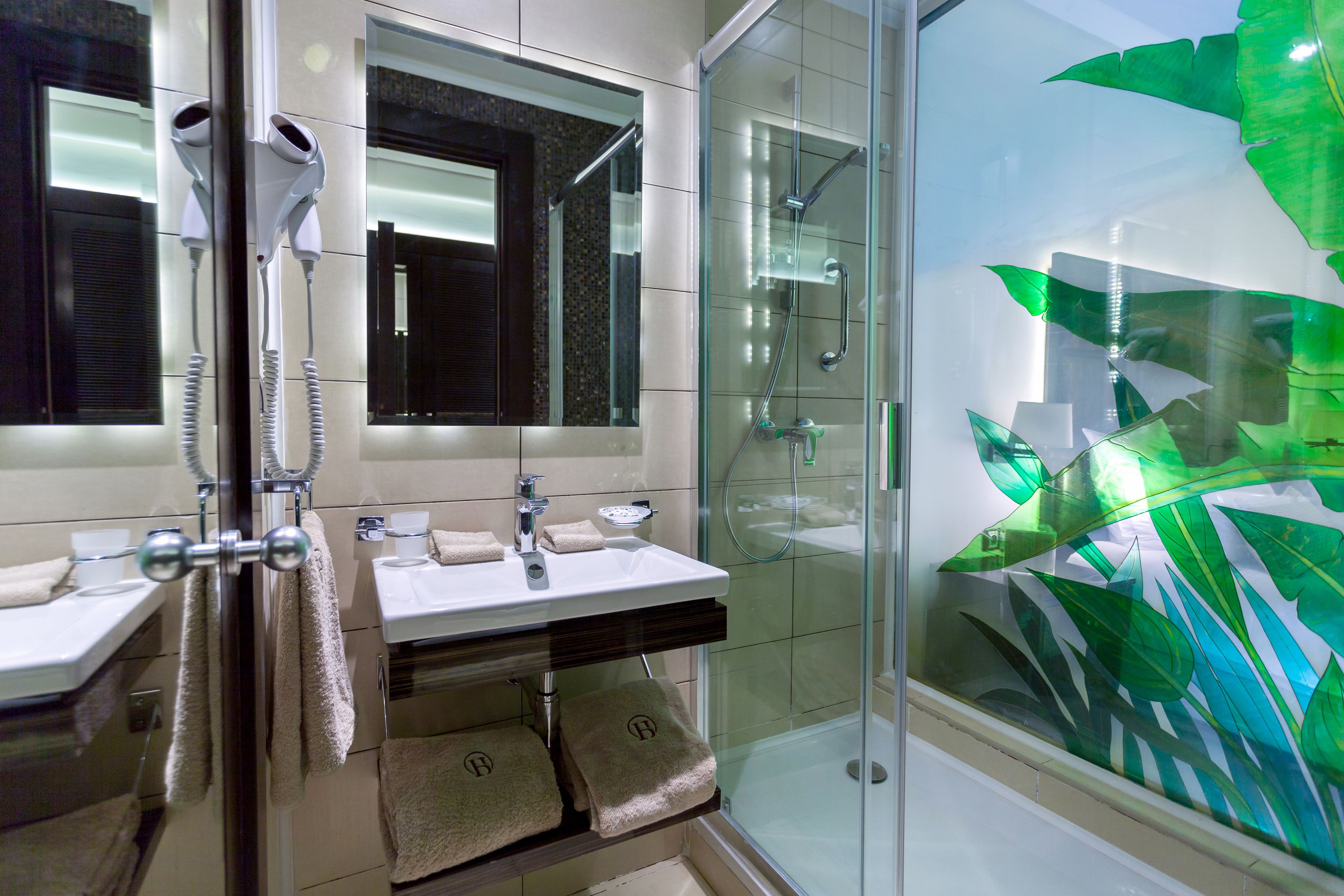 Un baño moderno con lavamanos dobles, secador de pelo, mampara de ducha de vidrio y pared decorativa de vidrio.