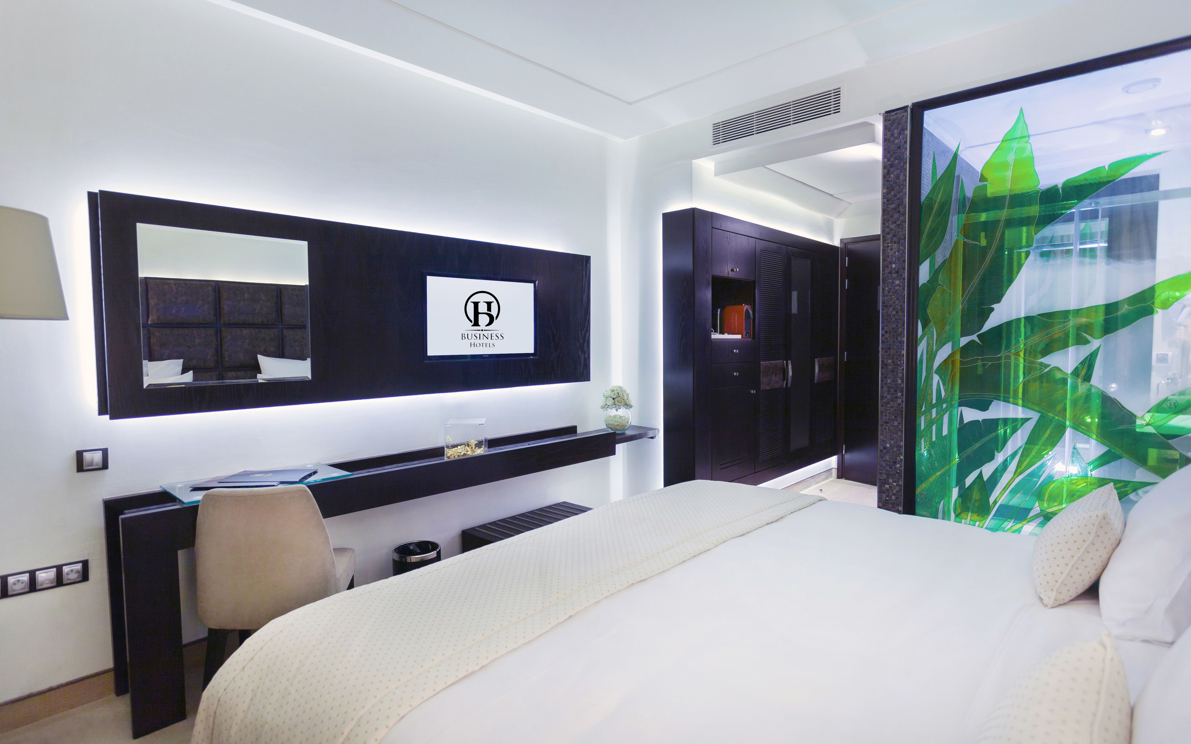 A modern hotel room with a large bed, a desk with a computer, a mirror, and a view of greenery through a window.