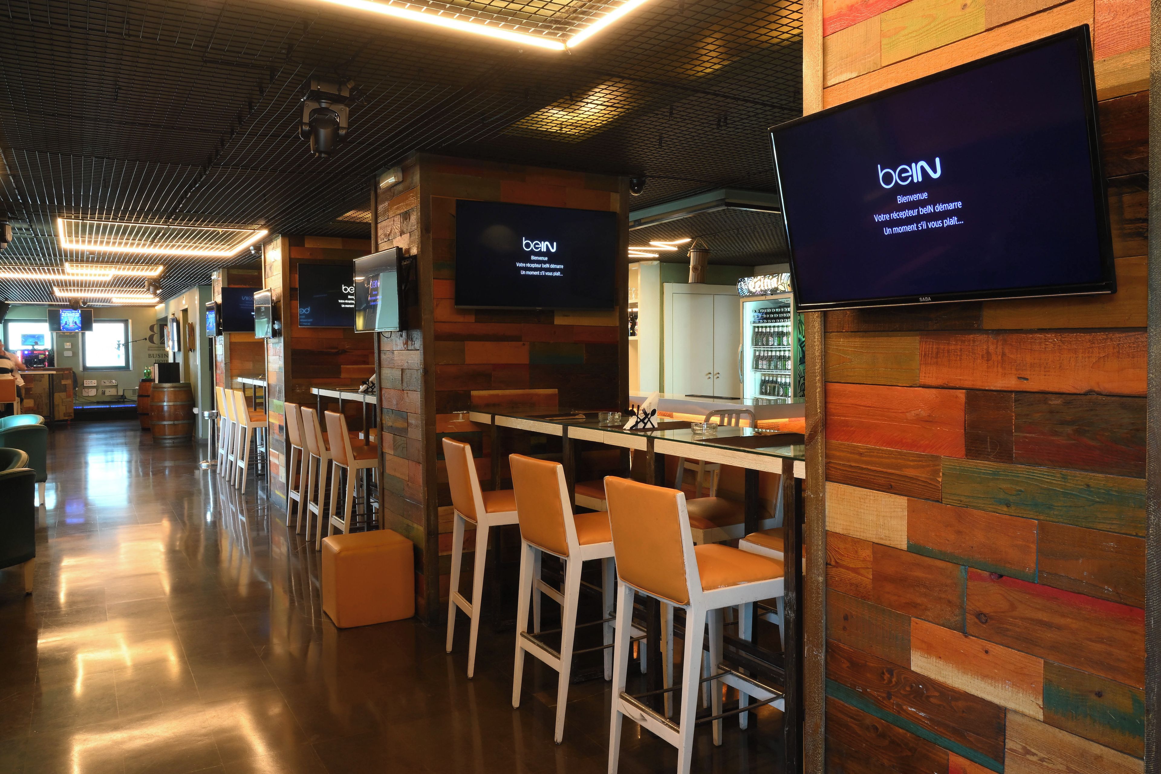 A modern bar area with wooden panel walls, bar stools, and a flat-screen TV displaying a welcome message.