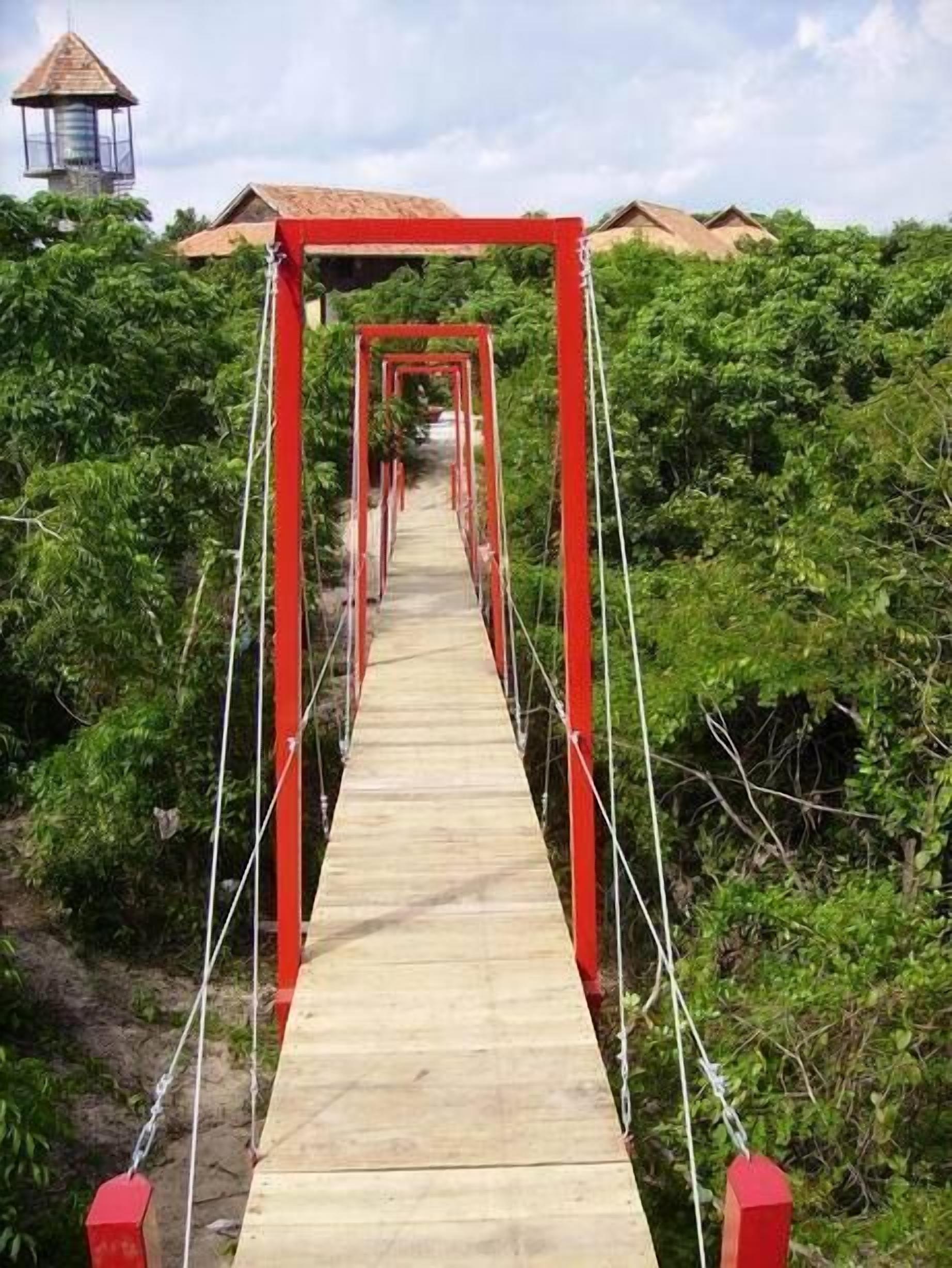 A wooden suspension bridge with red railings, surrounded by dense greenery.