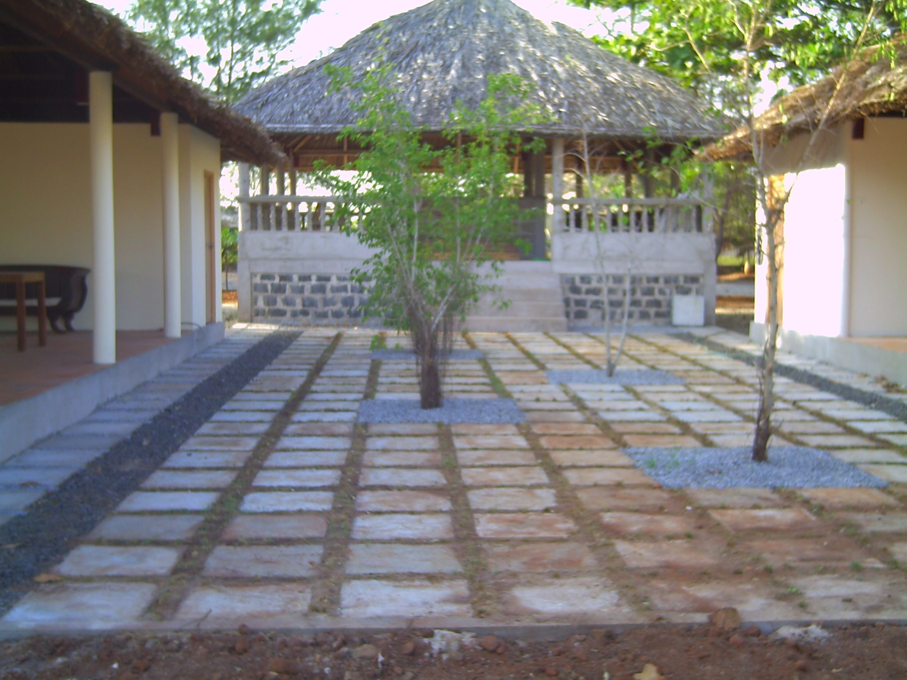 A courtyard with a thatched-roof gazebo, stone pathway, and a small tree.