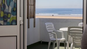 Ein Balkon mit weißen Plastikstühlen und einem Tisch, der auf einen Strand mit Menschen in der Ferne blickt.