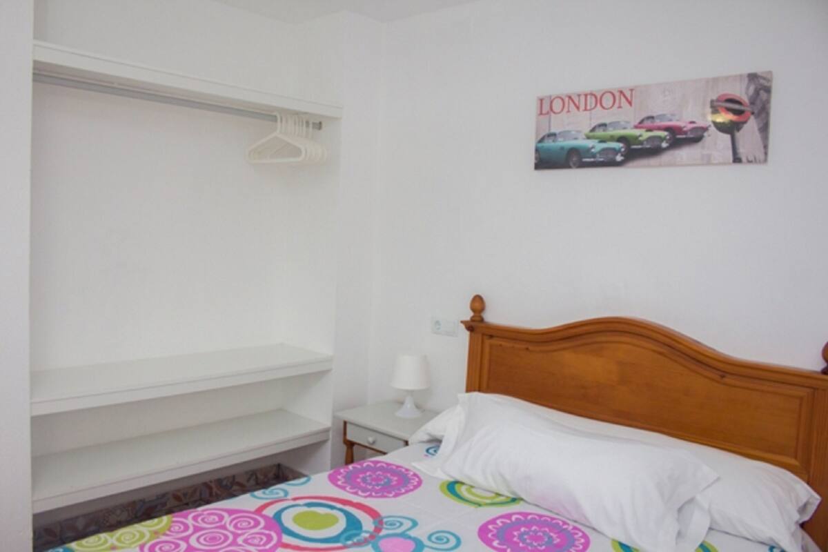 Une chambre avec une tête de lit en bois, un lit recouvert d’une courtepointe aux motifs circulaires et colorés, un oreiller blanc, une table de chevet avec une lampe et un tableau accroché au mur représentant des voitures, avec le mot « LONDRES ».