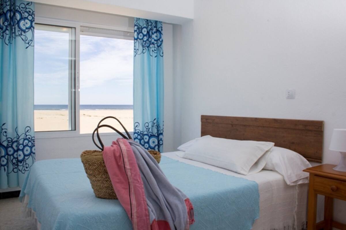 Une chambre à coucher avec un lit, une tête de lit en bois, une table de chevet avec une lampe, et une vue sur la plage par une fenêtre ornée de rideaux bleus.
