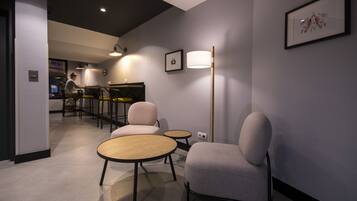 A modern interior with a seating area featuring round tables and cushioned chairs, a bar area in the background, and a framed picture on the wall.