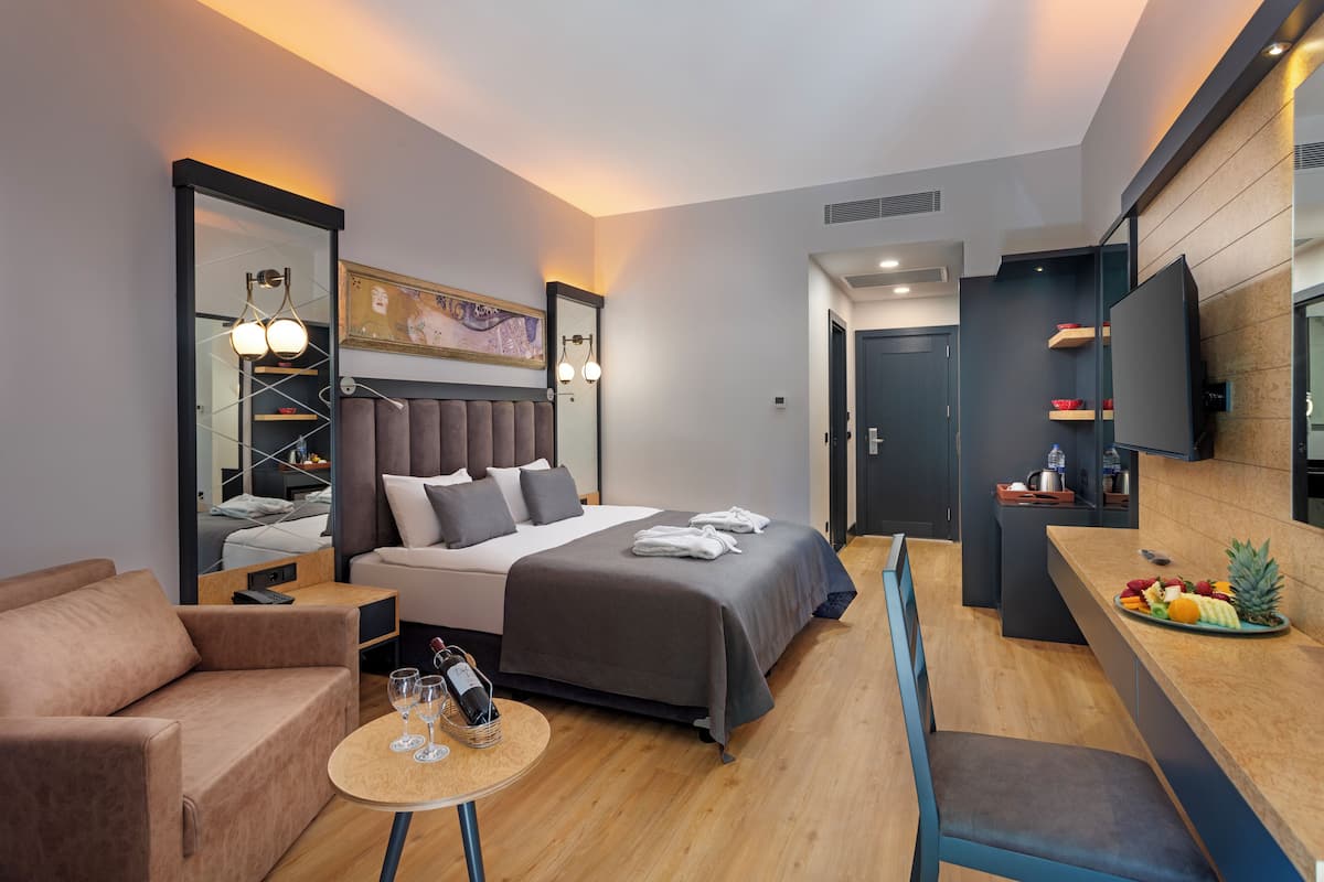 A modern hotel room with a bed, a sofa, a small table with glasses, a mirror, and a fruit tray on the counter.