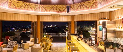 Zona de bar moderna con vistas a la ciudad de noche, con mobiliario de madera y techo con diseño.