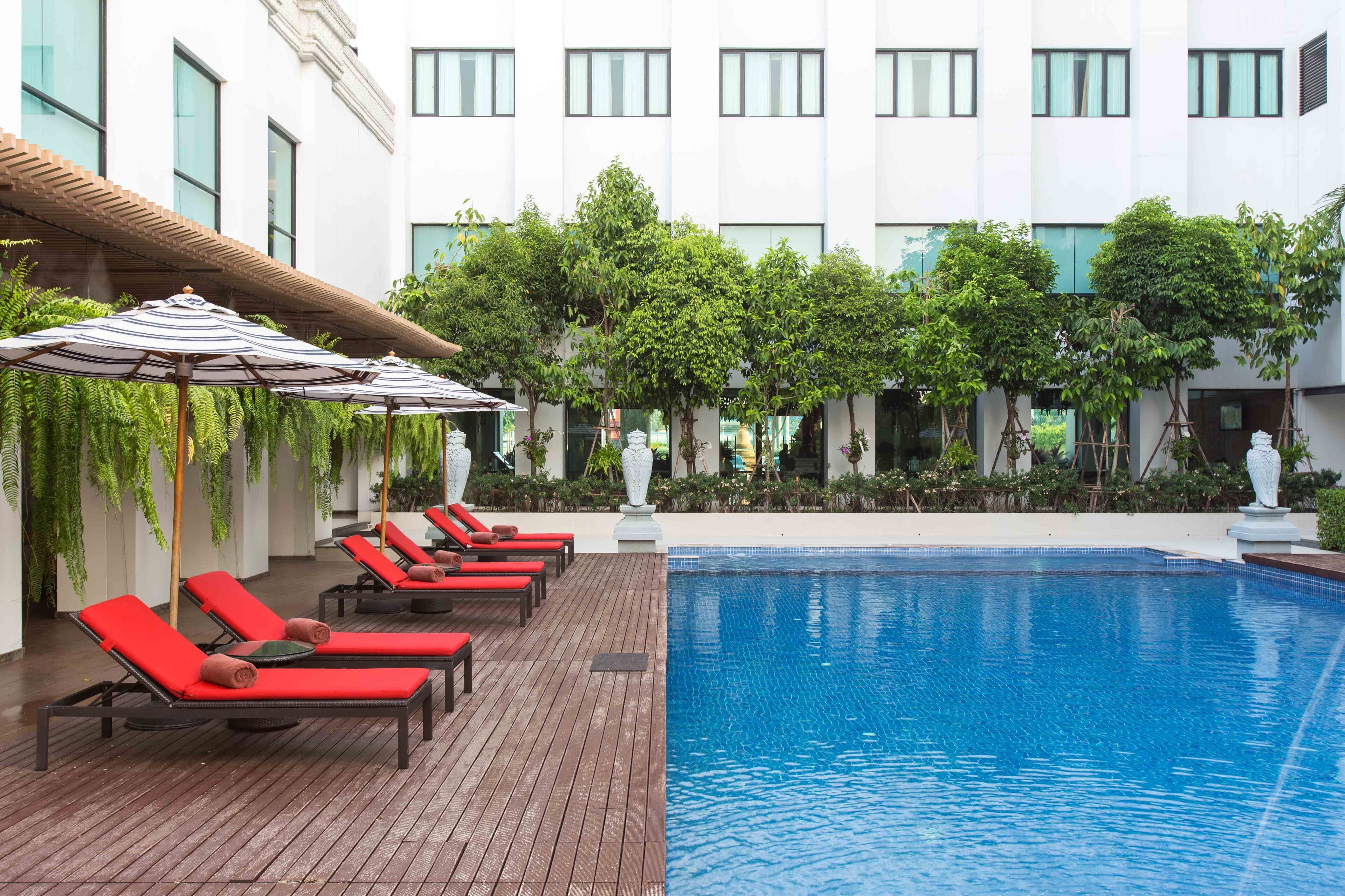 Zona de piscina con sillones de descanso y sombrillas, rodeada de follaje y una fachada de edificio moderna.