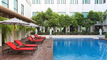Zona de piscina con sillones de descanso y sombrillas, rodeada de follaje y una fachada de edificio moderna.