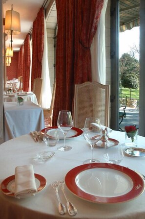 A dining table set with red and white plates, silverware, and wine glasses, with a view of a garden through large windows. - Domaine d'Auriac (Carcassonne)
