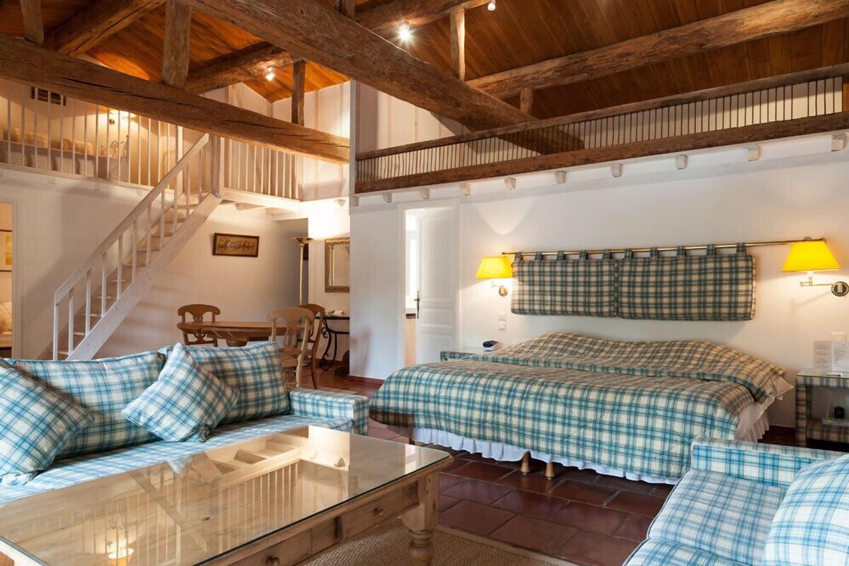 A cozy room with a wooden ceiling, a bed with plaid bedding, a glass table, and a staircase.