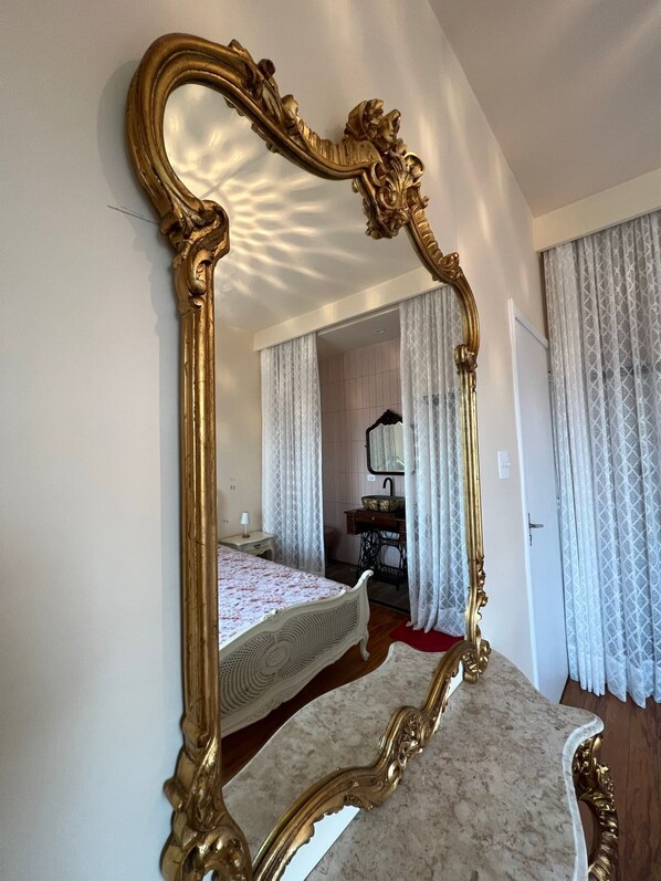 A bedroom with a large ornate mirror, a bed with a floral bedspread, a dresser, and a vanity with a mirror. - Paradiso Hostel (São Paulo)