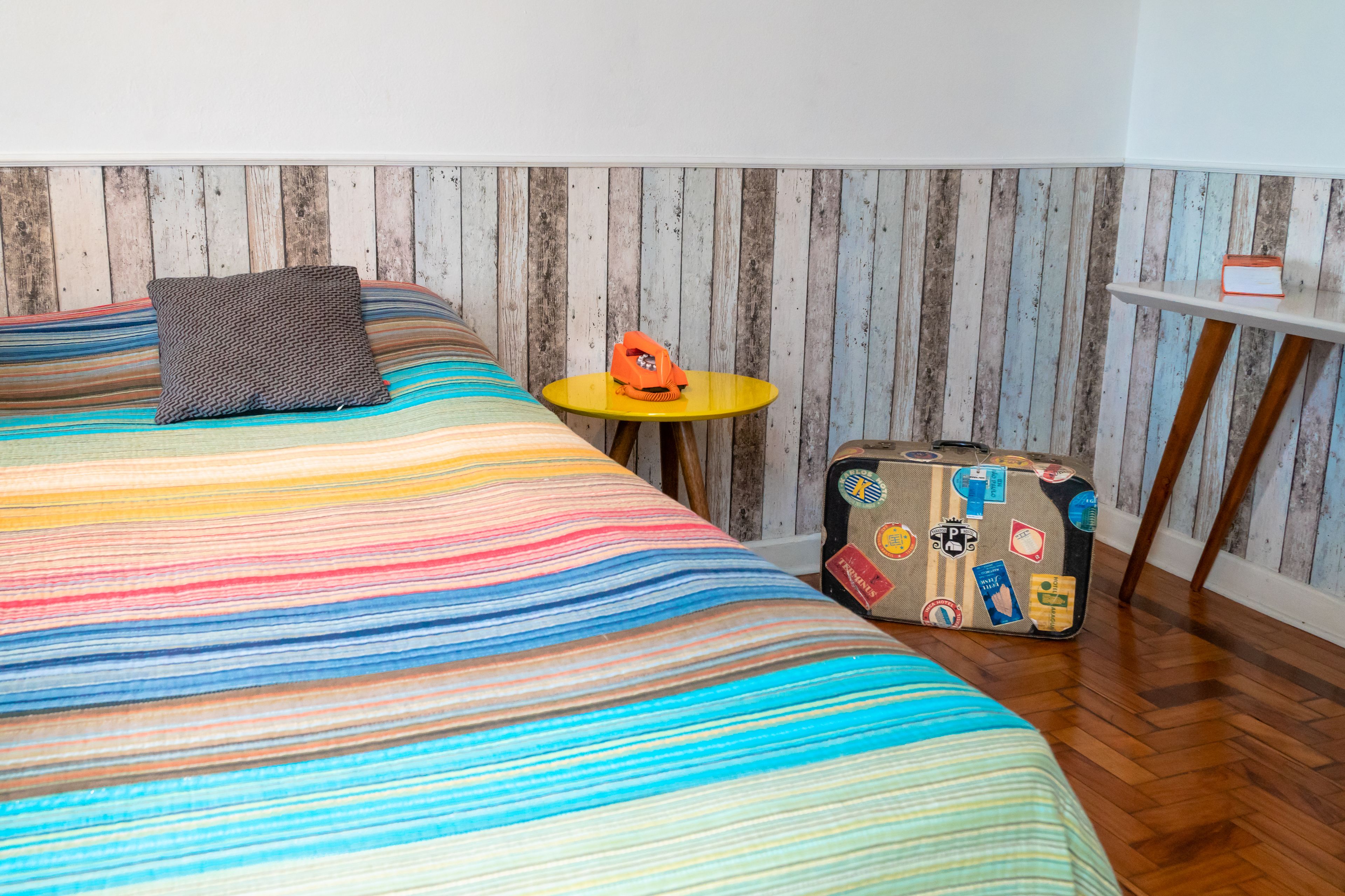 Un letto con una coperta a righe colorate, un tavolino con una valigia e una parete in legno a listelli verticali.