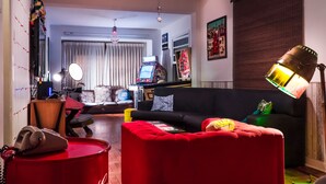 Una sala de estar con un sofá negro, un reposapiés rojo y un escritorio con una computadora. Hay una ventana con cortinas y un ventilador de techo.